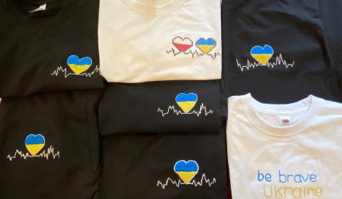 na zdjęciu są czarne i białe koszulki z logotypem w kształcie serca. W środku logotypu kolory Ukrainy i Polski