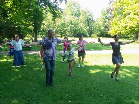 Na zdjęciu w parku widać kilku seniorów oraz kilkuletnią dziewczynkę, którzy ustawieni w 3 rzędach wykonują ruchy taneczne