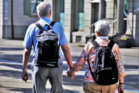 Na zdjęciu 2 osoby. Para seniorów w podróży z plecakami podróżnymi. Trzymają się za ręce spacerując po mięście. Przed soba mają ulicę.  