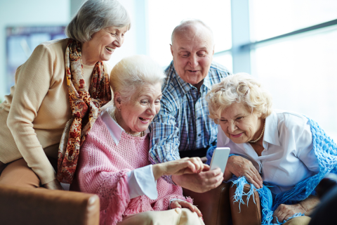 Zdjęcie przedstawia czwórkę seniorów, którzy patrzą wspólnie na telefon komórkowy. Są uśmiechnięci. Jedna osoba siedzi na środku pokoju w fotelu, reszta ją otacza. W tle widać rozjaśnione okna pomieszczenia. 