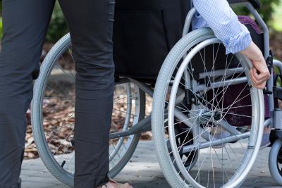 Na zdjęciu widać kadr osoby niepełnosprawnej na wózku i osobę pomagającą pchać wózek