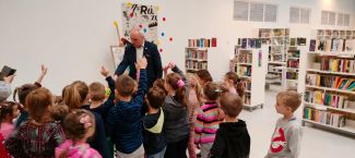 Na zdjęciu grupa dzieci i Pan Robert Pawliszko opowiadający dzieciom bajki w bibliotece. 