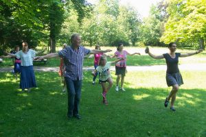 Na zdjęciu w parku widać kilku seniorów oraz kilkuletnią dziewczynkę, którzy ustawieni w 3 rzędach wykonują ruchy taneczne