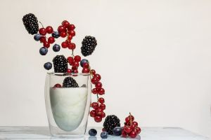 Na stole mamy szklankę wypełniona jogurtem i wpadające do niej owoce: czerwone porzeczki, jeżyny i jagody